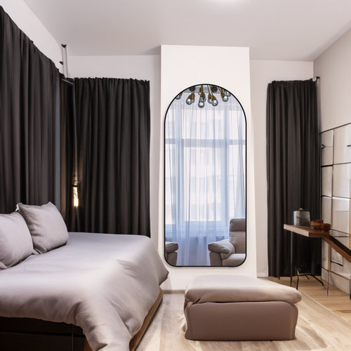 Full-length arched floor mirror with black metal frame, standing against a wall.