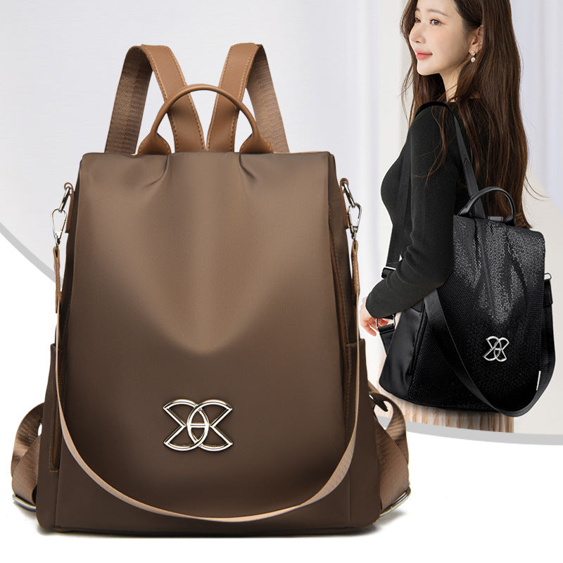 Brown backpack with curved front flap, gold logo emblem, top handles and shoulder straps
