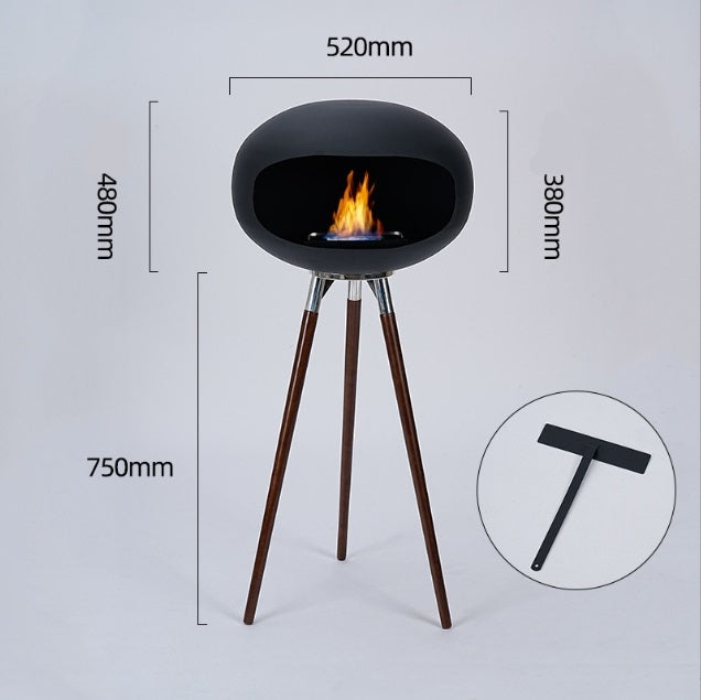 Black round ethanol fireplace bowl on three wood tripod legs, flame burning in the tray.